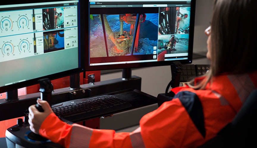 A woman in orange work wear sitting at a desk in a control room supervising drilling operations via computer screens. (photo)