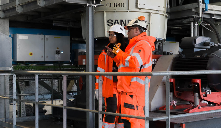 Två personer i orange arbetskläder omgivna av utrustning från Sandvik för krossning och sortering (foto)